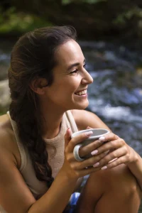 Femme souriante et confiante prenant un café en pleine nature, illustrant la détente et le bien-être, symbolisant l'importance de prendre soin de soi dans le cadre des services de coaching de MouvZone.