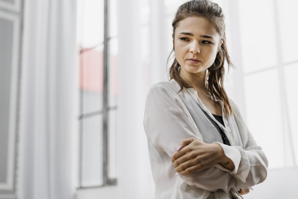 Femme debout, bras croisés et épaules rentrées, qui doute d'elle dès que c'est pour elle et pas pour les autres